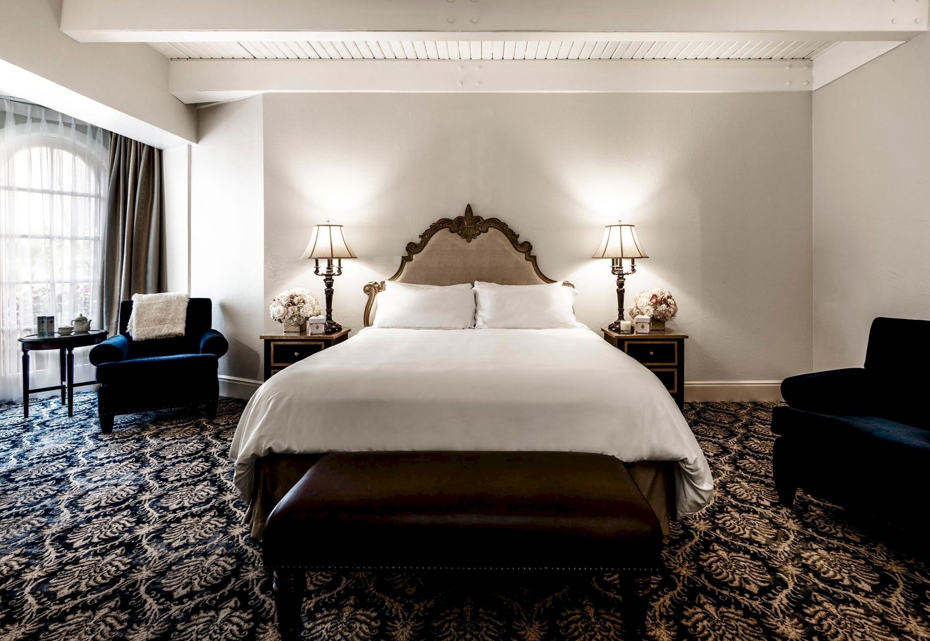 A luxurious hotel bedroom with a carved headboard bed, two lamps, nightstands, curtains, and a patterned carpet.