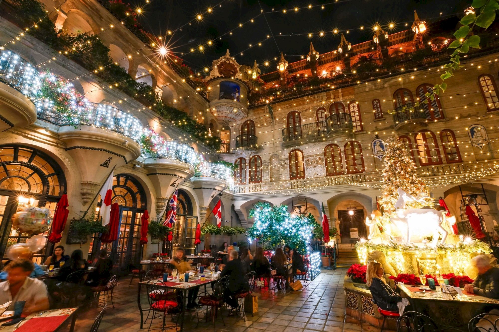A grand courtyard dressed for Christmas with strings of lights, flags, a sculpture display, and guests dining at night.