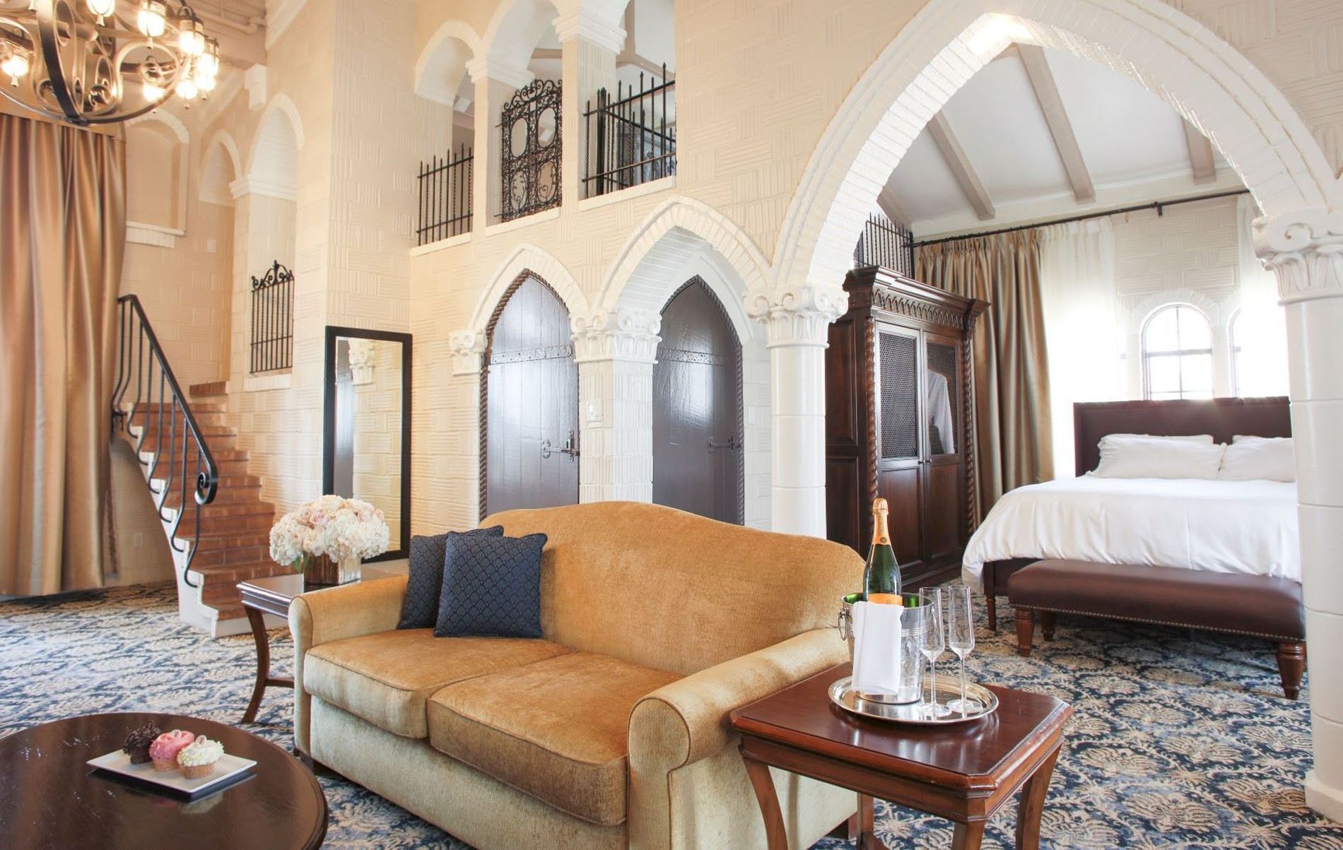 A luxurious hotel suite interior with an arched white ceiling, couch, coffee table, sparkling drinks, and a grand bed.