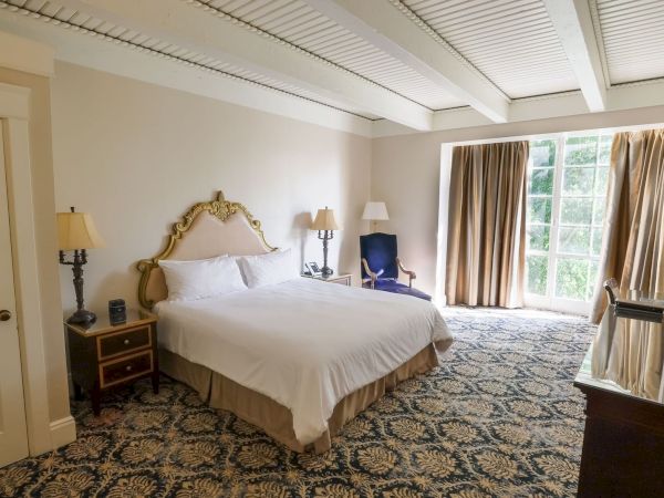 A tidy bedroom with a large bed, lamps, curtains, carpet, and an office chair by the window.