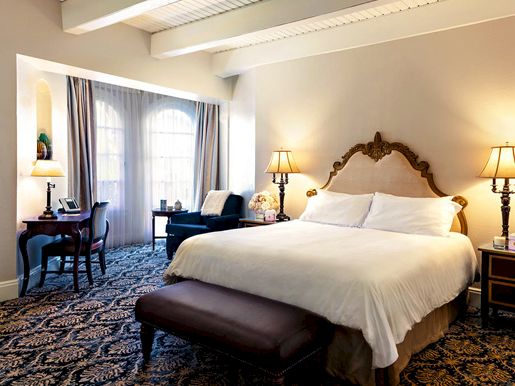 A bright hotel bedroom with a large bed, bedside lamps, curtains, and a desk chair beside the window.