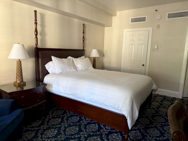 A tidy hotel-style bedroom with a made bed, two lamps, dark headboard, and a closed white door in the background.