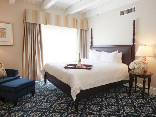 A neatly made hotel bedroom with a king bed, curtains, side table lamp, and flower arrangement.