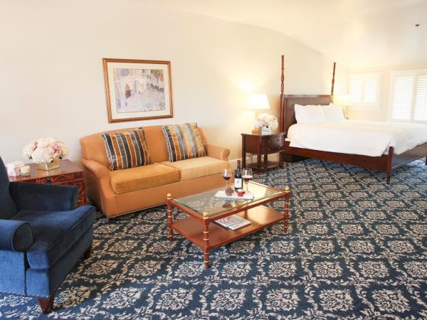 A bright hotel suite with a sofa, coffee table, and a neatly made bed by windows.