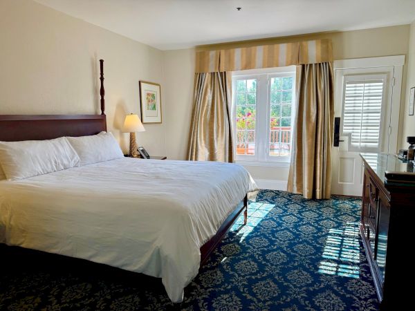 A tidy hotel bedroom with a large bed, bedside lamp and phone, striped curtains, blue patterned carpet, and windows.