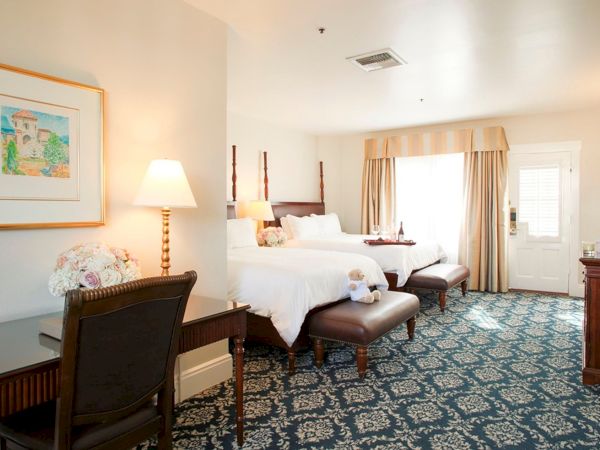 A bright hotel bedroom with twin beds, patterned carpet, curtains, bedside lamps, a dresser, and a framed wall picture.