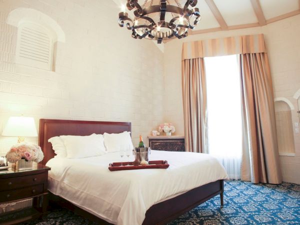 Bright hotel bedroom with a king bed, ornate chandelier, and a champagne tray by the window behind flowing curtains.
