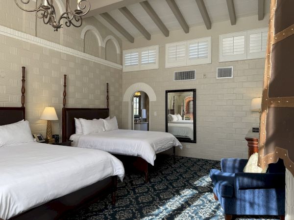 A modern hotel room with two beds, a mirrored wall, dark carpet, white brick walls, and a chandelier above.