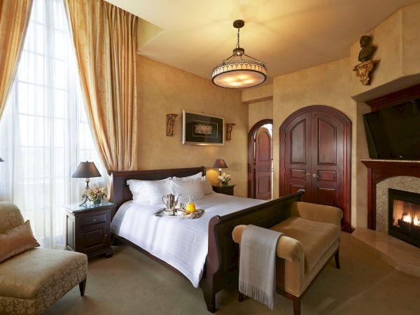 A luxurious hotel bedroom with a four-poster bed, arched doors, chandelier, armchairs, and a lit fireplace.