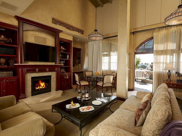 A cozy luxury living room with a fireplace, TV wall, dining table, and large arched windows with curtains.