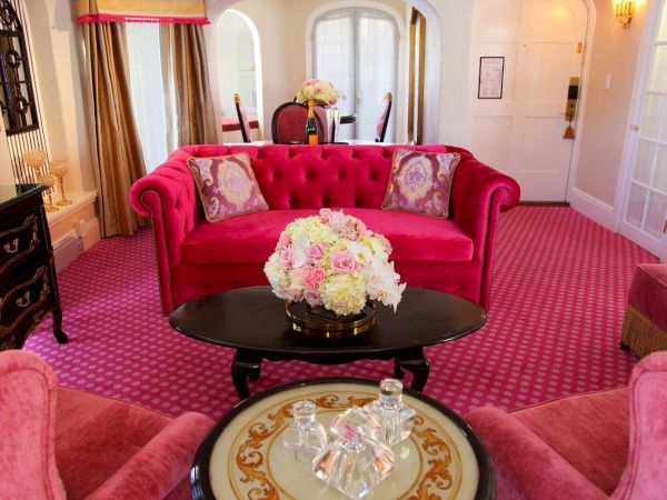 A stylish lounge with a bright pink tufted sofa, ornate curtains, floral centerpiece, and bottles set on a coffee table.