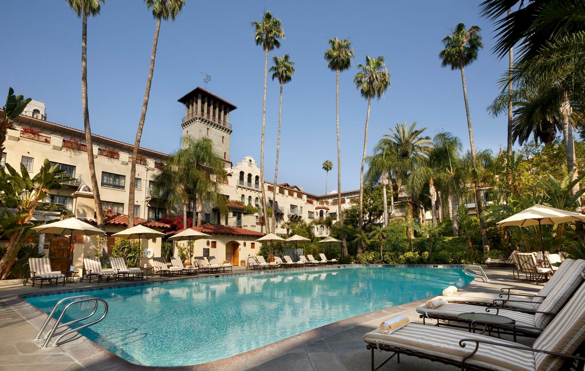 A sunny hotel courtyard with a blue swimming pool, striped loungers, umbrellas, and tall palm trees.