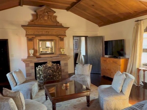 Elegant living room with plush sofas, an ornate fireplace, a coffee table, wood beams overhead, and a TV in the background.
