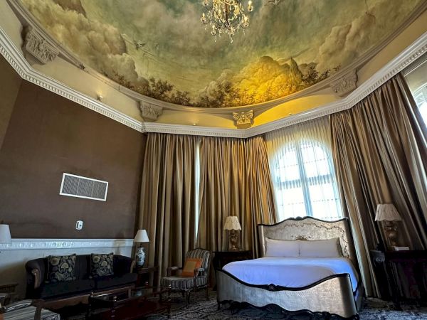 An elegant hotel room with a chandelier, painted ceiling mural, gold curtains, and a bed with sofa in view.