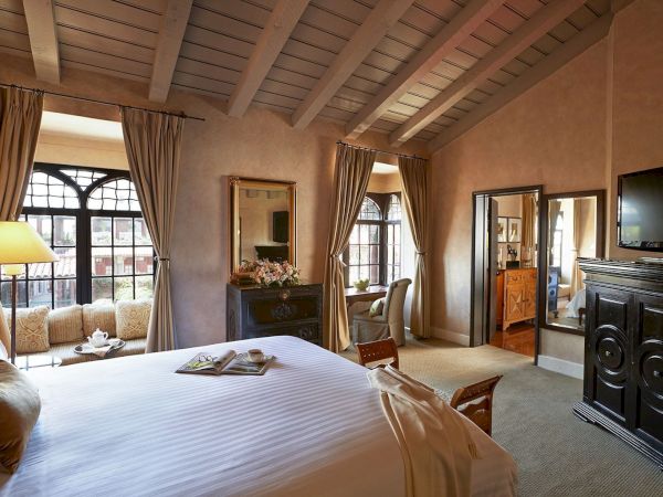 A cozy hotel suite with a large bed, tall curtains, warm decor, and a dark fireplace in the corner.