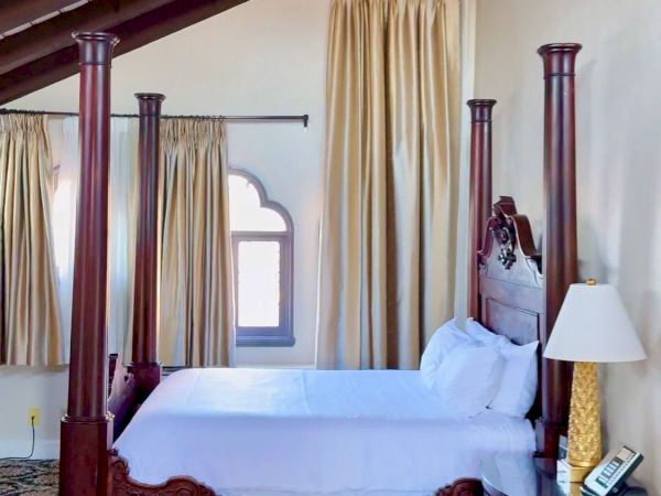 A four-poster bed with tall curtains under wood beams, flanked by matching posts, with a bedside lamp and carpeted floor.