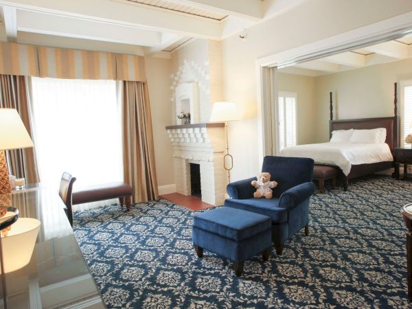 Bright hotel suite with a blue sofa, fireplace, and a bed by large windows, with patterned carpet throughout.