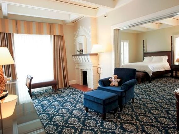 A cozy hotel suite living room with armchairs, an ottoman, lamp, fireplace, patterned carpet, and a teddy bear.