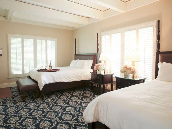 A bright bedroom with two beds, dark wooden headboards, bedside lamps, and large white-shutter windows.