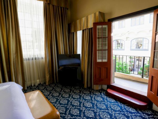 A hotel room with a bed, TV, dark curtains, and an open door to a balcony garden view.