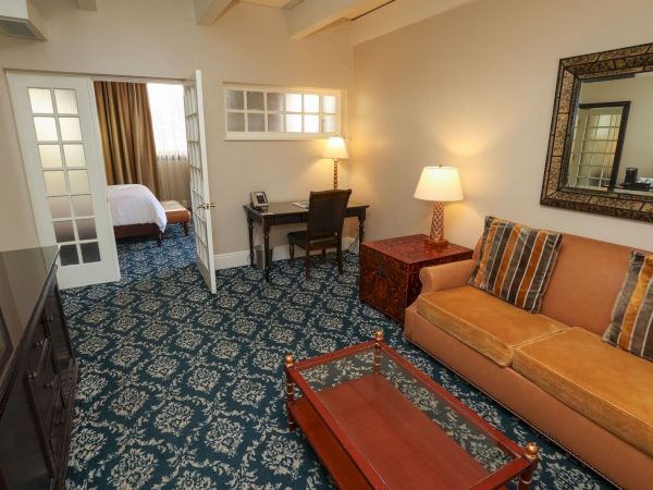 A cozy hotel sitting room with a tan sofa, patterned blue carpet, lamps, a small desk, and a large wall mirror.