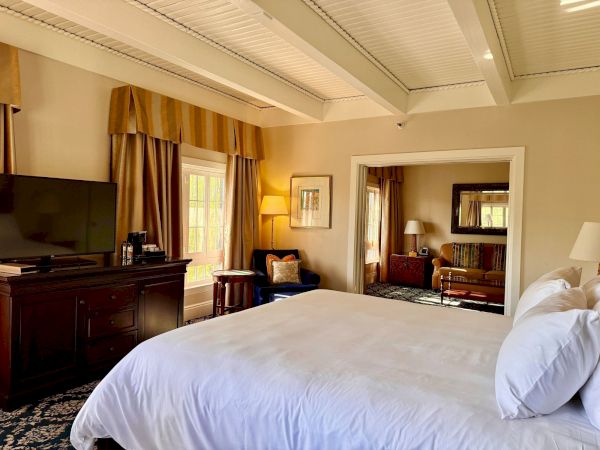 A cozy hotel room with a large bed, TV and dresser, warm curtains, and a seating area with lamps and mirrors.
