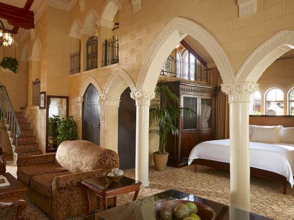 Luxurious hotel suite with cream arches, ornate columns, a king bed, and upholstered seating in a warm lounge.