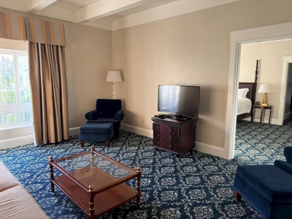 A hotel lounge/bedroom features a wall-mounted TV, dark armchair, floor lamp, and two beds in an adjoining doorway.