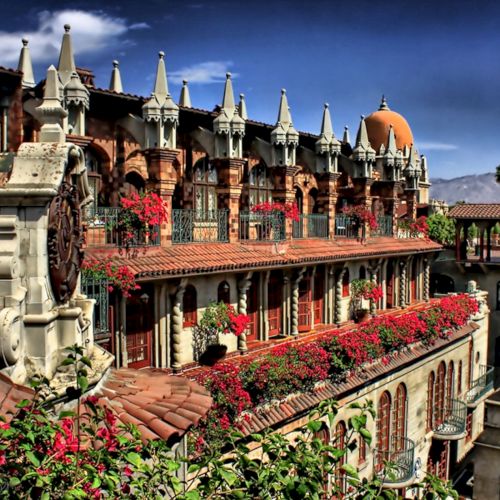 Ornate historic courtyard with brick arches, white spires, and lots of red flowers under a bright blue sky.