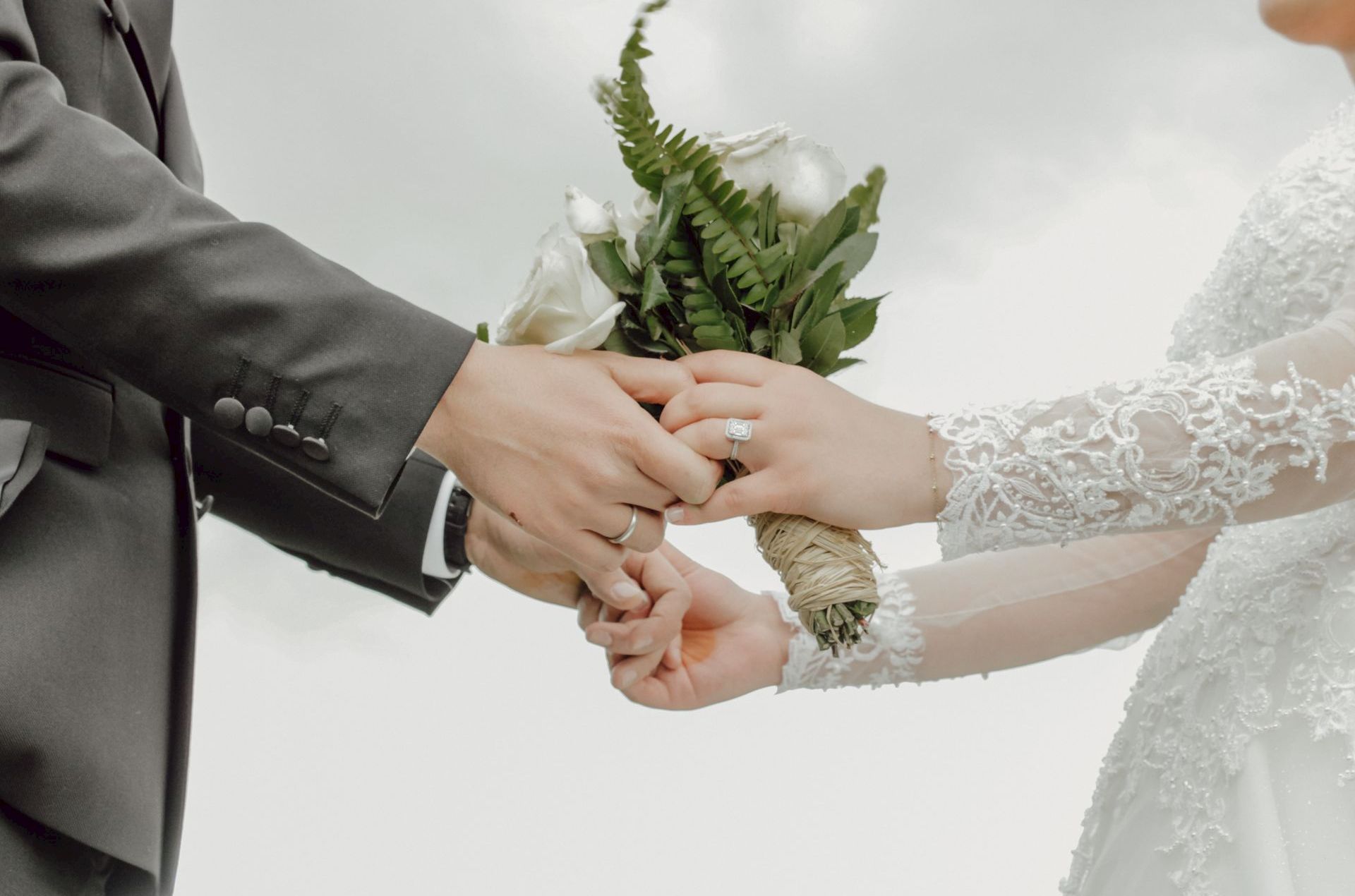 A groom in a suit and a bride in lace hold a bouquet while showing wedding rings in a tender, wedding-day moment.
