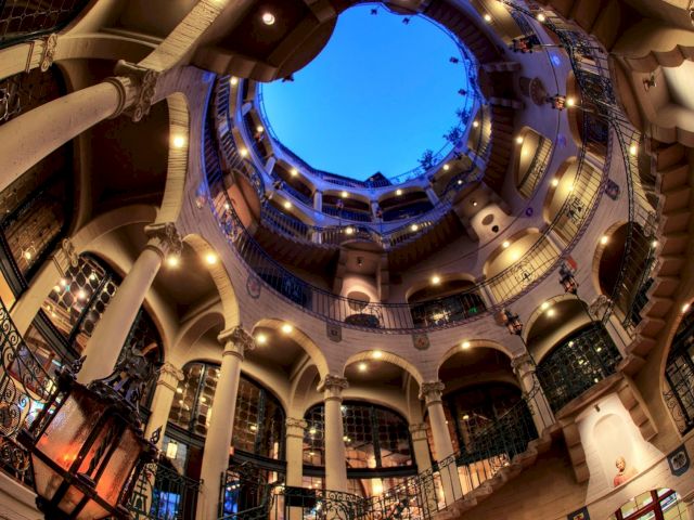 A grand indoor courtyard with curved balconies and arches around a glowing circular skylight, lit in blue.