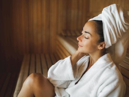 Relaxing woman in a spa setting wrapped in a white towel, eyes closed, leaning with her head supported by her hand.