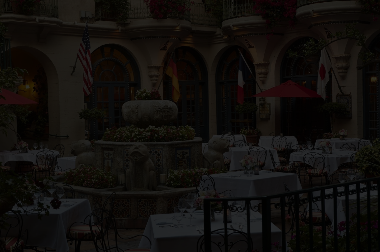 An elegant outdoor courtyard set for dining with white-clothed tables, flower planters, and national flags hanging on arched walls.