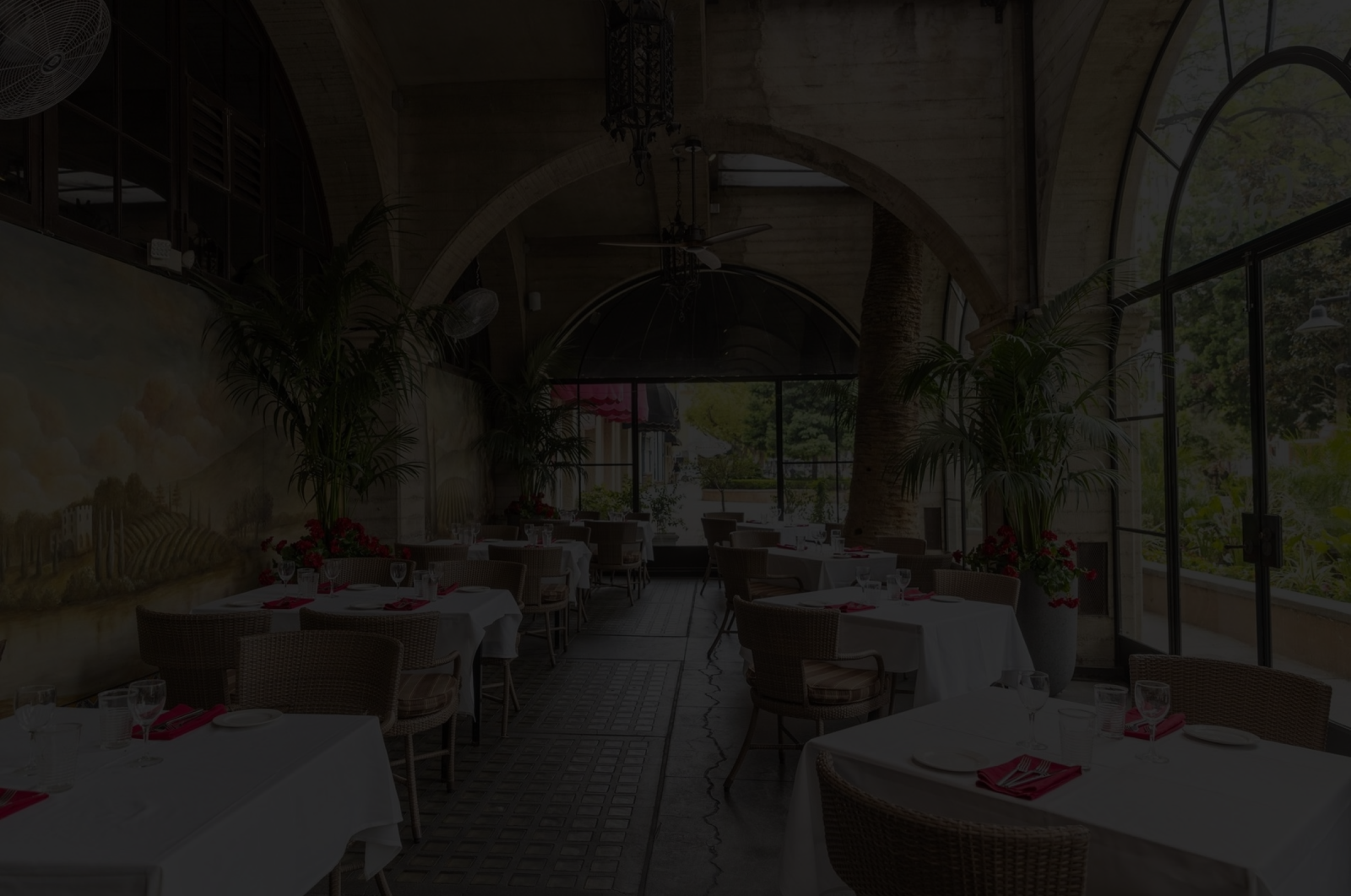 An elegant dining patio with banquet tables, chairs, potted palms, and stone arches under warm indoor light.