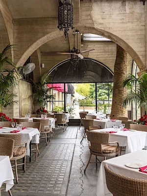A bright indoor patio dining room with white table settings, red accents, tropical plants, arched stone walls, and a ceiling fan.