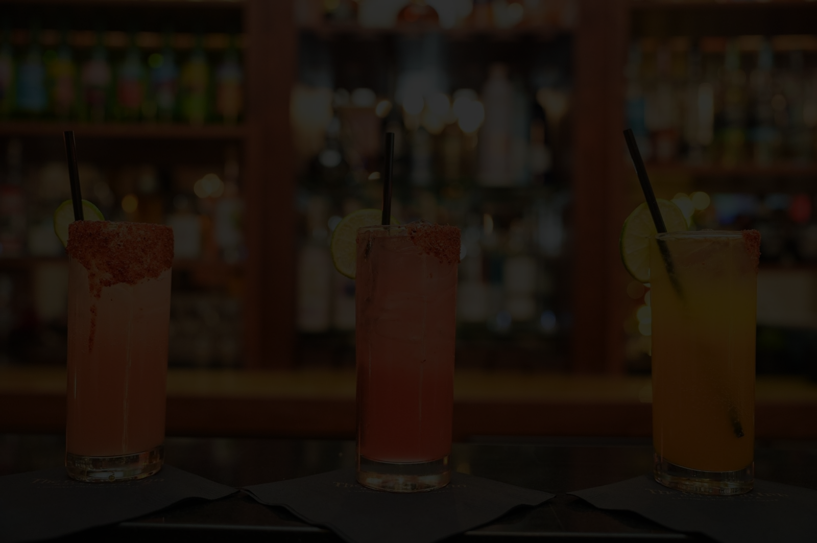 Three colorful drinks in tall glasses with straws&mdash;one pinkish-red, one pale, and one bright yellow&mdash;served on a bar counter.