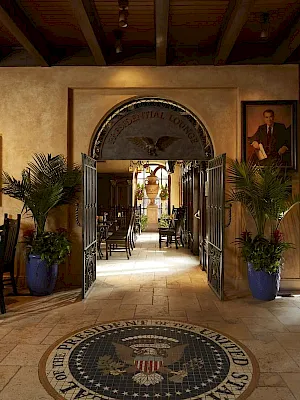 Interior lounge hallway with framed portraits, potted palms, iron gates, and a large eagle emblem rug on the floor.
