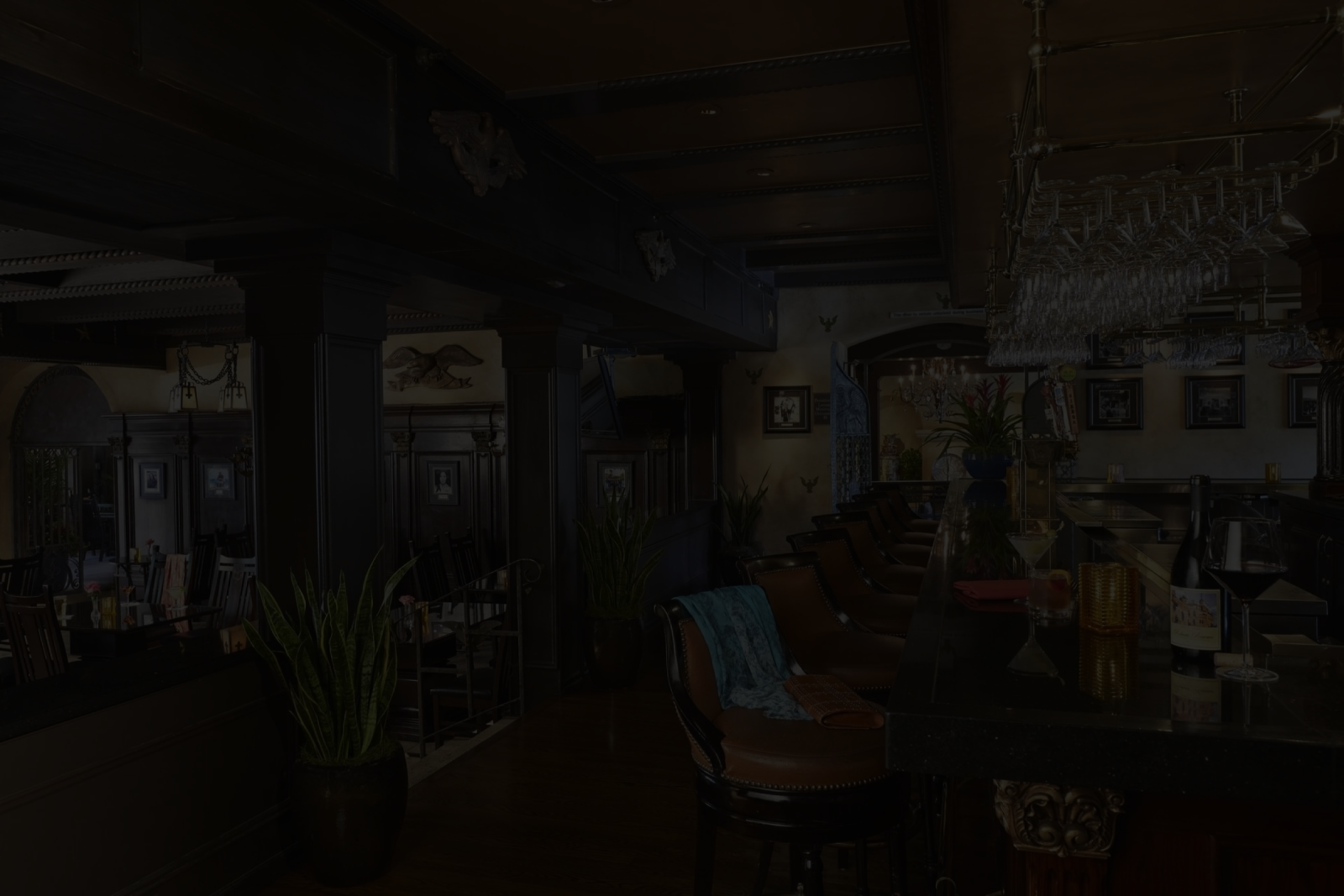 A dimly lit lounge/kitchen with dark beams, exposed pipes, framed pictures, chairs, plants, and glassware.