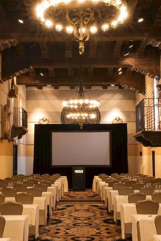 An elegant banquet hall with candlelit chandeliers, long dining tables, and religious murals lining the walls.