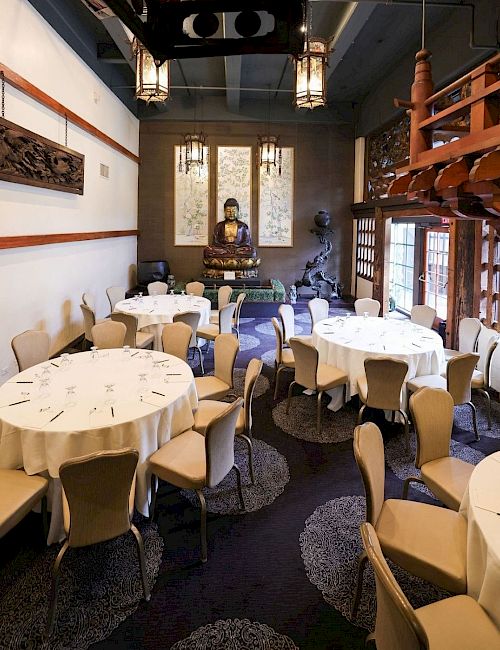 A classy dining hall with neatly set round tables, warm hanging lantern lights, and a central Buddha statue in a calm, ornate room.