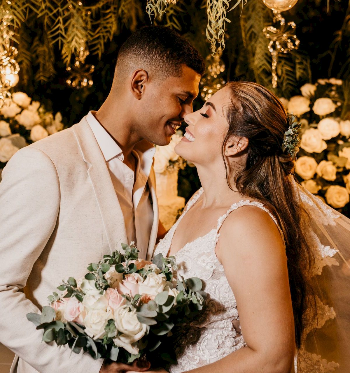 A bride and groom in wedding attire share a tender kiss under warm lights and floral decor, smiling in celebration.
