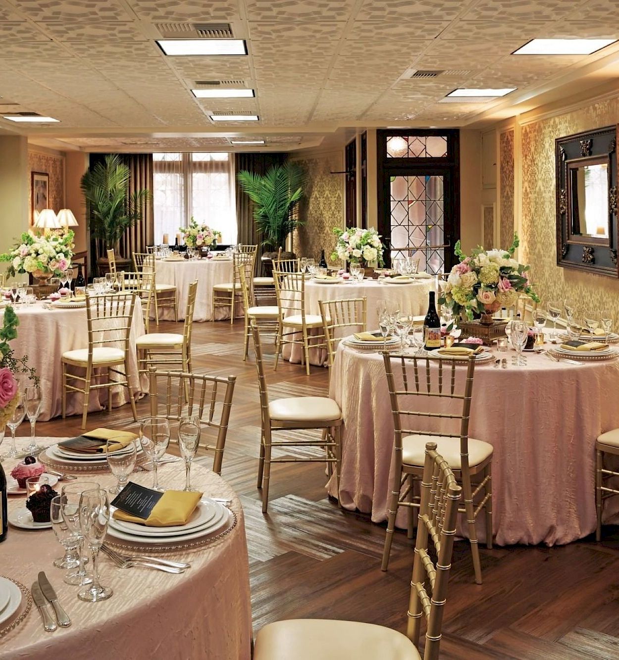 Elegant event hall set for a wedding or banquet with round tables, floral centerpieces, candles, and gold chiavari chairs.