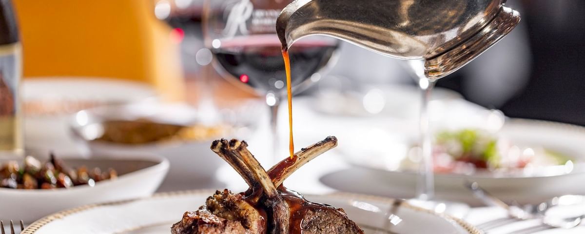 A chef pours rich sauce onto grilled lamb chops on a plated dish, with silverware nearby, in a restaurant setting.