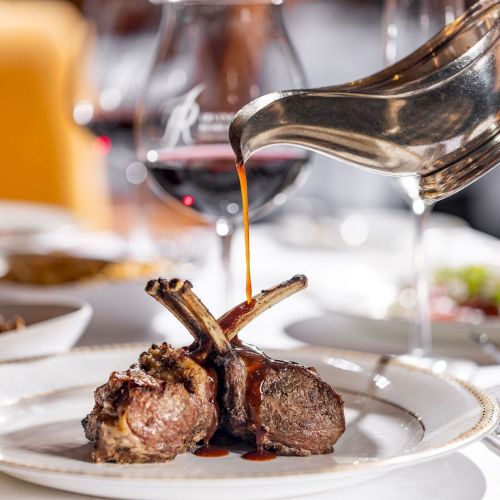 A chef pours rich sauce from a pan onto plated grilled lamb chops, finishing a restaurant-style dish with gravy drizzling down.