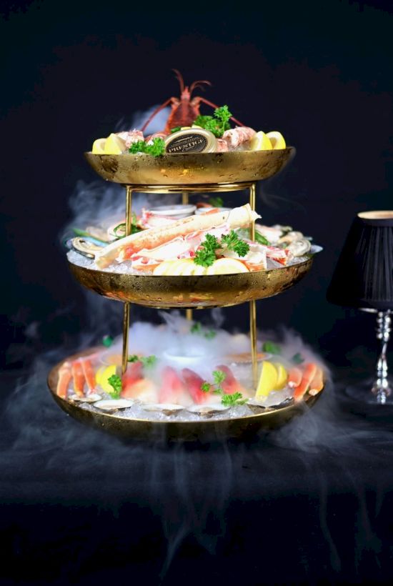 A sizzling seafood platter in a golden two-tier chafing stand, topped with crab, lemon, herbs, and steam.