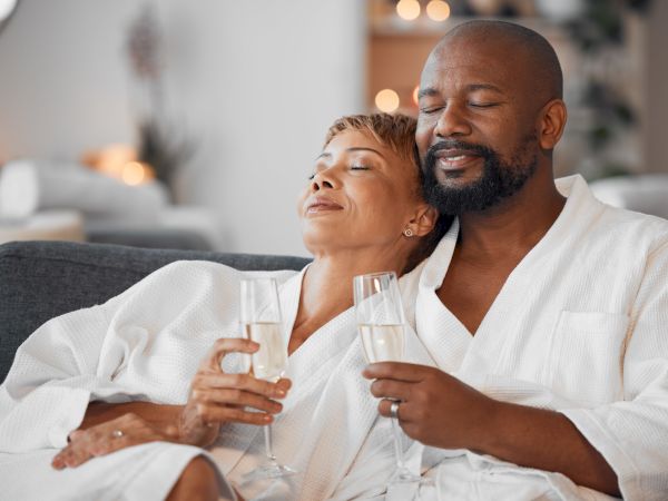 A smiling couple in white robes clinks champagne glasses, cuddled on a couch during a cozy celebration.