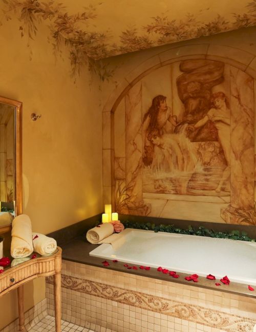 A romantic spa bathroom with a large white bathtub, candles, rose petals, and a wall mural of classical figures.