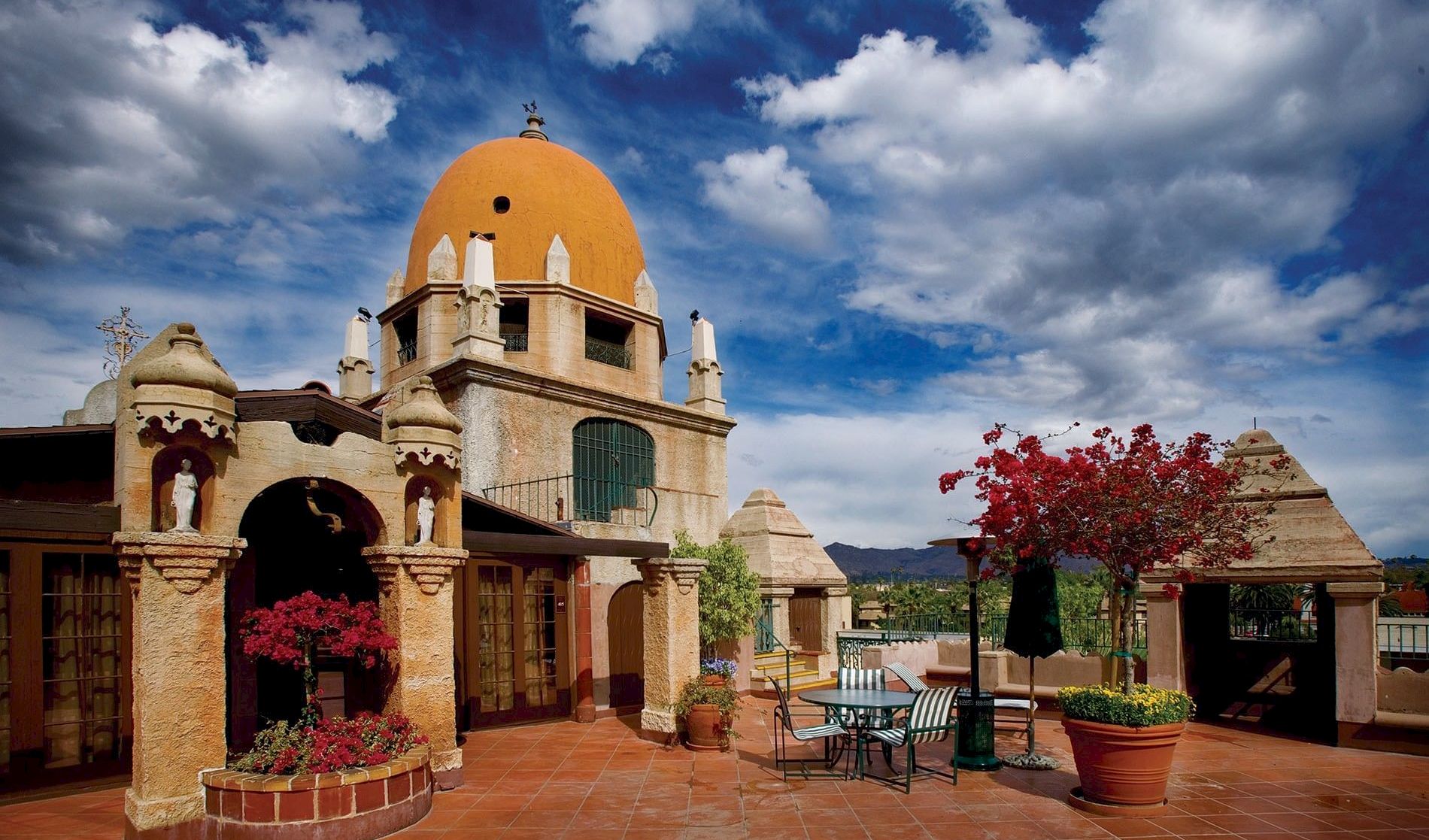 Yellow-domed church over a sunny courtyard with arches, potted flowers, and patio furniture under dramatic blue clouds.
