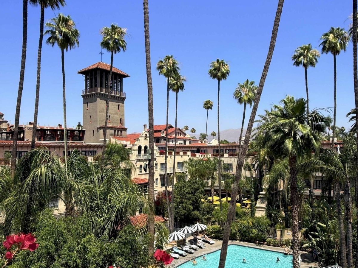 A sunny tropical resort scene with a turquoise swimming pool, palm trees, umbrellas, and a small castle-like tower building.