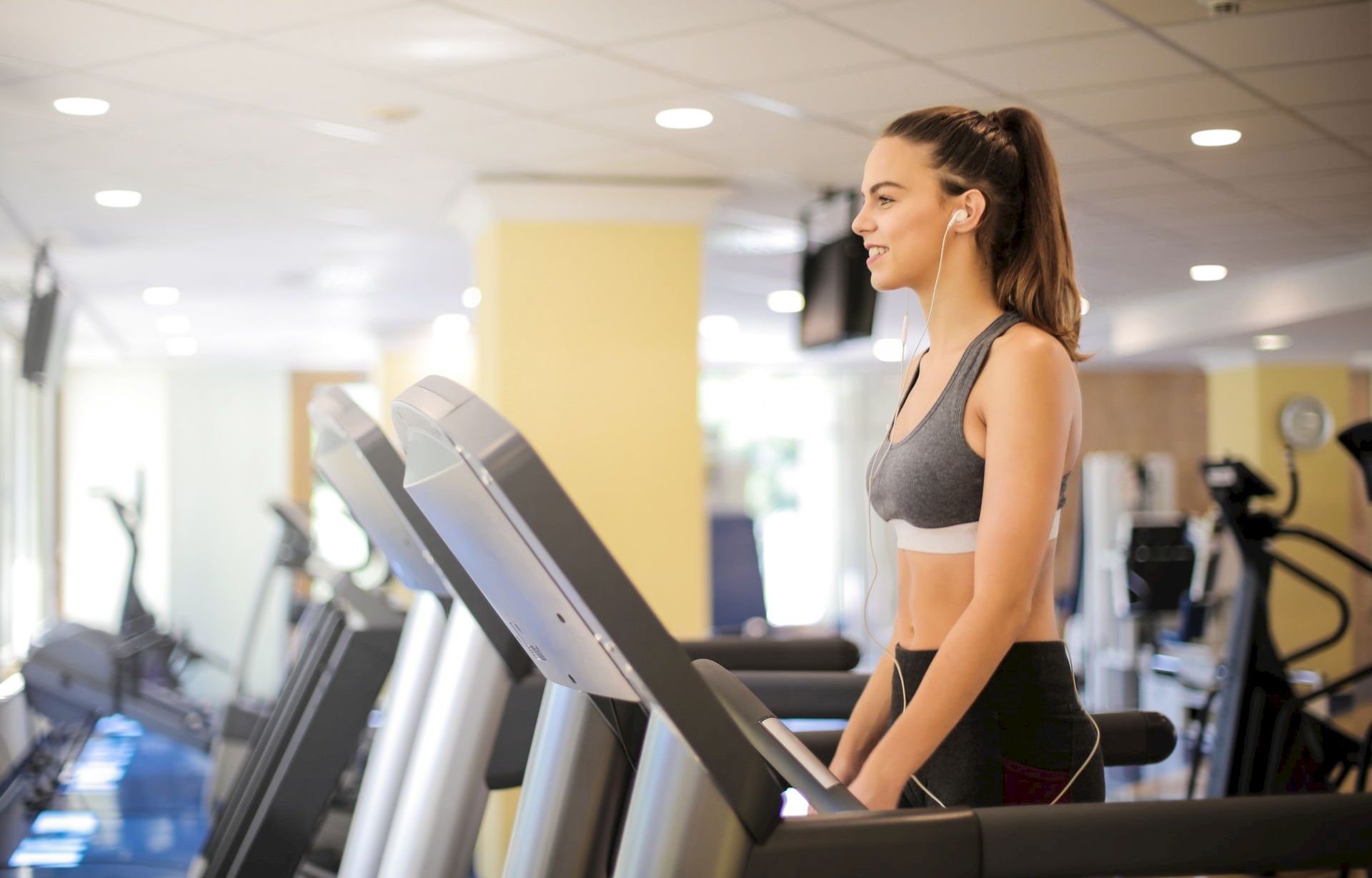 A woman works out on a treadmill in a gym, wearing earbuds and looking focused while running.